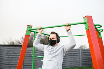 A young man is working on a horizontal bar outdoors during quarantine, he is doing pull-ups in medical mask on the face. Healthy lifestyle concept