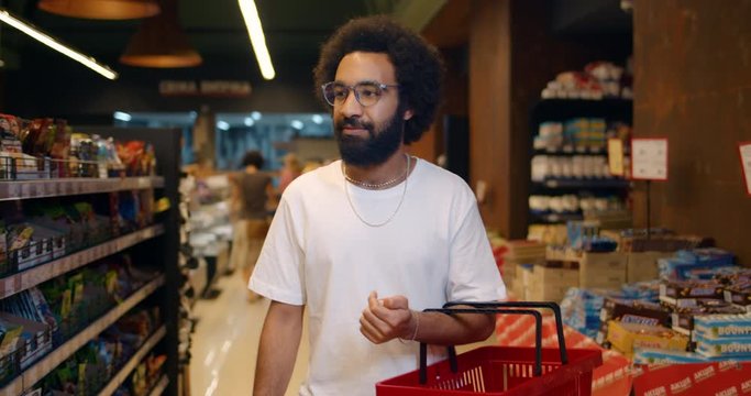 Front view of guy in glasses walking through store section with shopping basket and looking around. Handsome bearded man in 30s wearing casual clothes and buying food in grocery shop