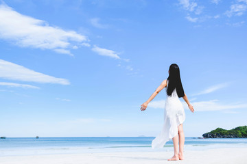 Asian woman, long black hair, wore white dress and hat standing at the beach,arms open and facing back by the sea with copy space of blue sky summer holiday and vacation concept.