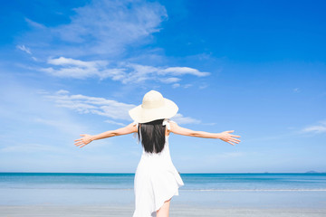 Fototapeta na wymiar Asian woman, long black hair, wore white dress and hat standing at the beach,arms open and facing back by the sea with copy space of blue sky summer holiday and vacation concept.