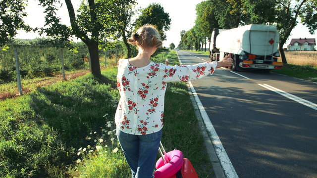 Traveler woman hitchhiking on a sunny road and walking. Young happy backpacker woman looking for a ride to start a journey on a sunlit country road.