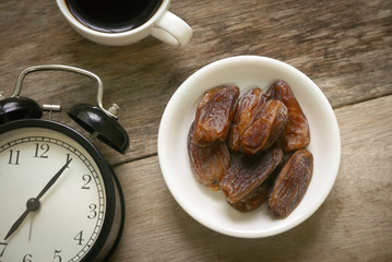 Selective focus and top view of a dried dates fruit in a white bowl together with a cup of coffee and vintage clock on wooden background. Ramadan holy month for muslim concept where they are fasting.