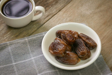 Selective focus of a dried dates fruit in a white bowl together with a cup of coffee on wooden background. Ramadan holy month for muslim concept where they are fasting.