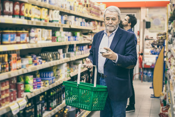 Mature man shopping in grocery store. Serious man holding shopping basket and choosing goods in supermarket. Shopping concept