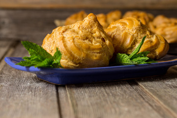 Delicious big cream puffs with cream in hand painted blue oval saucer with mint leaves on aged wooden background. Selective focus.