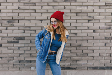 Fototapeta na wymiar Wonderful white lady in red hat happy dancing on the street. Outdoor photo of pretty blonde girl in denim clothes having fun on brick background.