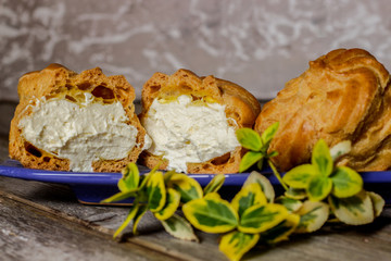 Delicious big cream puffs with cream in hand painted blue oval saucer with mint leaves on aged wooden background. Selective focus.