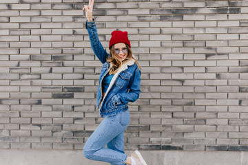 Slim girl in trendy jeans having fun on the street in cold spring day. Blissful female model in denim attire dancing on urban background and waving hands.