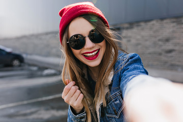 Laughing young woman in casual red hat making selfie. Outdoor portrait of magnificent girl posing on urban background with sincere smile.