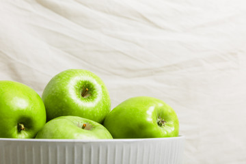 Green apples in a bowl on a light fabric background. Harvest apples, summer season, fresh fruits. Copy space.