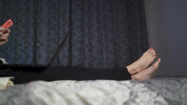 Young woman with bare feet and red nail polish lying in bed smiling using her smartphone