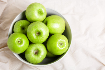 Green apples in a bowl on a light fabric background. Harvest apples, summer season, fresh fruits. Top view.