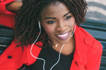 Smiling young lady with earphones relaxing on street. Beautiful African American woman with dreadlocks listening to music. Leisure concept