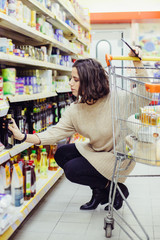 Woman holding smartphone and choosing goods in store. Young woman leaning at shopping trolley, holding cell phone and buying products in supermarket. Shopping concept