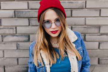 Close-up portrait of ecstatic european woman wears stylish red hat. Attractive young lady in denim outfit standing in confident pose near brick background.