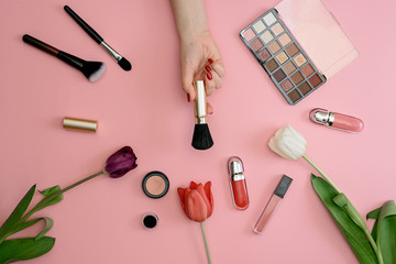 Girl holds makeup brush set of cosmetics and flowers on pink background