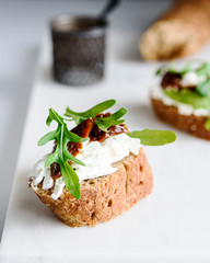 Italian bruschetta (sandwich, crostini, toast) with confit tomatoes, feta, olive oil and arugula leaves on white marble background. Traditional italian appetizer or snack, antipasto. Selective focus