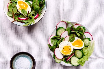 Radish, cucumber, herb mix and soft-boiled egg salad in two bowls on white tablecloth. Healthy nutrition. Selective focus