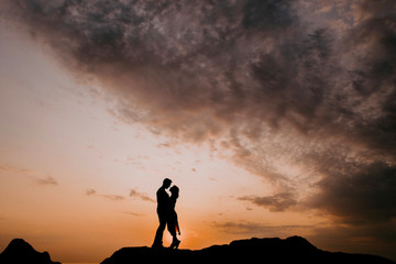 Silhouette of a couple in love against the background of dawn. A guy and a girl stand on stones by the sea at sunrise