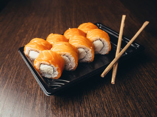 Food delivery, Philadelphia rolls in a black box with chopsticks. Brown wood background.