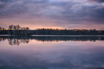 Faraway dark forest reflecting in the lake level during cloudy sunset in dark violet colors. The lake Sadská near to czech city Nymburk.