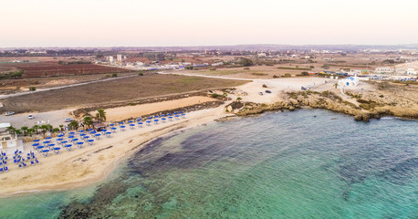 Aerial bird's eye view of coastline sunset, traditional landmark white and blue chapel at Agia Thekla beach, Ayia Napa, Famagusta, Cyprus from above.Tourist attraction golden sand bay, church, sunbeds