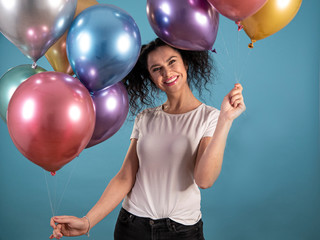 Portrait of young woman with black curly hair in casual dress smiling and holding party balloons isolated on blue background