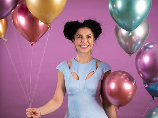 Portrait of young woman with black curly hair in blue party outfit and ballons smiling isolated on pink background