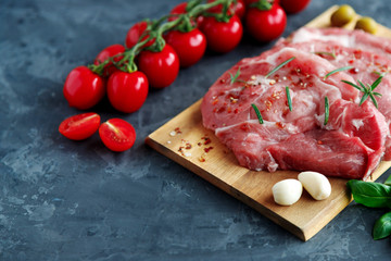 Fresh pork chops in spices on a wooden board, near cherry tomatoes on a branch and garlic cloves on a dark background. Place for text. Cooking meat for grilling, frying