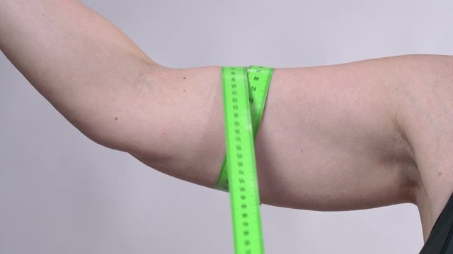 A middle-aged woman with saggy skin determines the circumference of her hand with a measuring tape, on a white background.