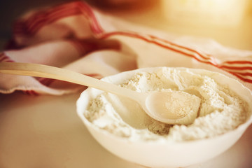 On a white table is a white bowl with crumbly flour and a wooden spoon to mix it, and next to it is a white fly with red stripes, illuminated by sunlight.