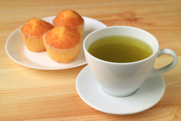 Cup of Hot Green Tea with Blurry Madeleine Cupcakes in Background Served on Wooden Table
