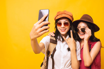 Two Asian pretty young girl smiling wear sunglasses and take a selfie by smartphone together, Young women backpacker take a selfie in studio yellow background.