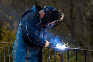 Fototapeta na wymiar A man in a blue jacket is engaged in welding. A welder in a black mask repairs an iron fence. A man at work.