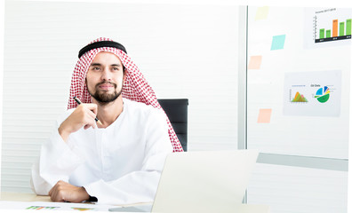 Portrait of Iranian in traditional muslim clothing sitting at workplace in front of computer and graph in board