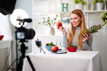 Food blogger cooking fresh vegan salad of fruits in kitchen studio, filming tutorial on camera for video channel. Female influencer holds apple, pineapple and talks about healthy eating. Fructorianism