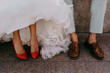 The feet of a wedding couple, the bride in bright red shoes, and the bride in brown shoes.