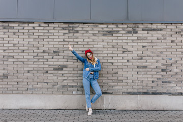 Amazing slim woman in blue jeans posing near brick wall in good spring day. Refined caucasian girl in denim jacket and white sneakers having fun on urban background.