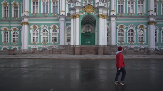 A hipster guy in a red jacket and burgundy hat is walking along the Hermitage.