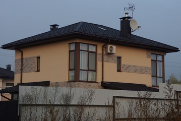 large brown private house with a window under a tiled roof behind a gray metal fence in dry grass against the sky