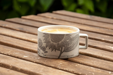 White matte cup with a monochrome pattern and with coffee on a wooden table on a blurred background of a green bush