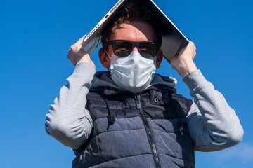 A young man in sunglasses and medical mask against blue sky covers his head from sun and viruses with modern laptop. Working remotely during coronavirus pandemic. Spring 2020. Man hiding under laptop