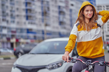 The girl rides a bicycle around the city and straightens the hood with his hand.