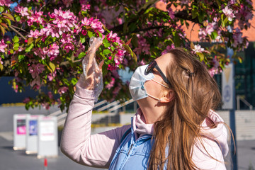 Girl or young woman in a medical mask and sunglasses sniffs flowers on blooming cherry or Apple...
