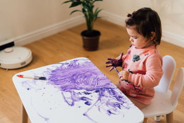Toddler girl painting white table with purple paint. Creative child mess. Homeschooling. 