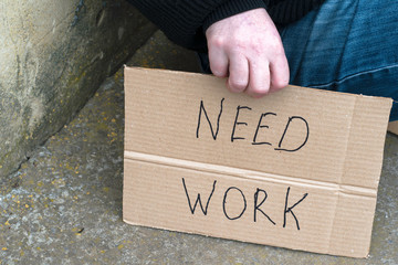 unemployed man sitting on the ground holding the cardboard sign saying need work