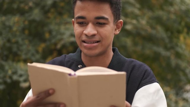Concentrated young african guy student sit on bench reading book outdoors.