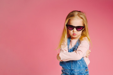 Fototapeta premium Young cute smiling girl in sunglasses is offended and angry on pink background. Concept of childhood.