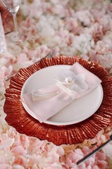 Wedding. Banquet. Glass table with pink and white flowers and red and white plates on it, served with cutlery, flowers and crockery.
