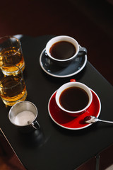Top view on two cups of coffee, sugar and 2 glasses of whiskey on the black wooden table